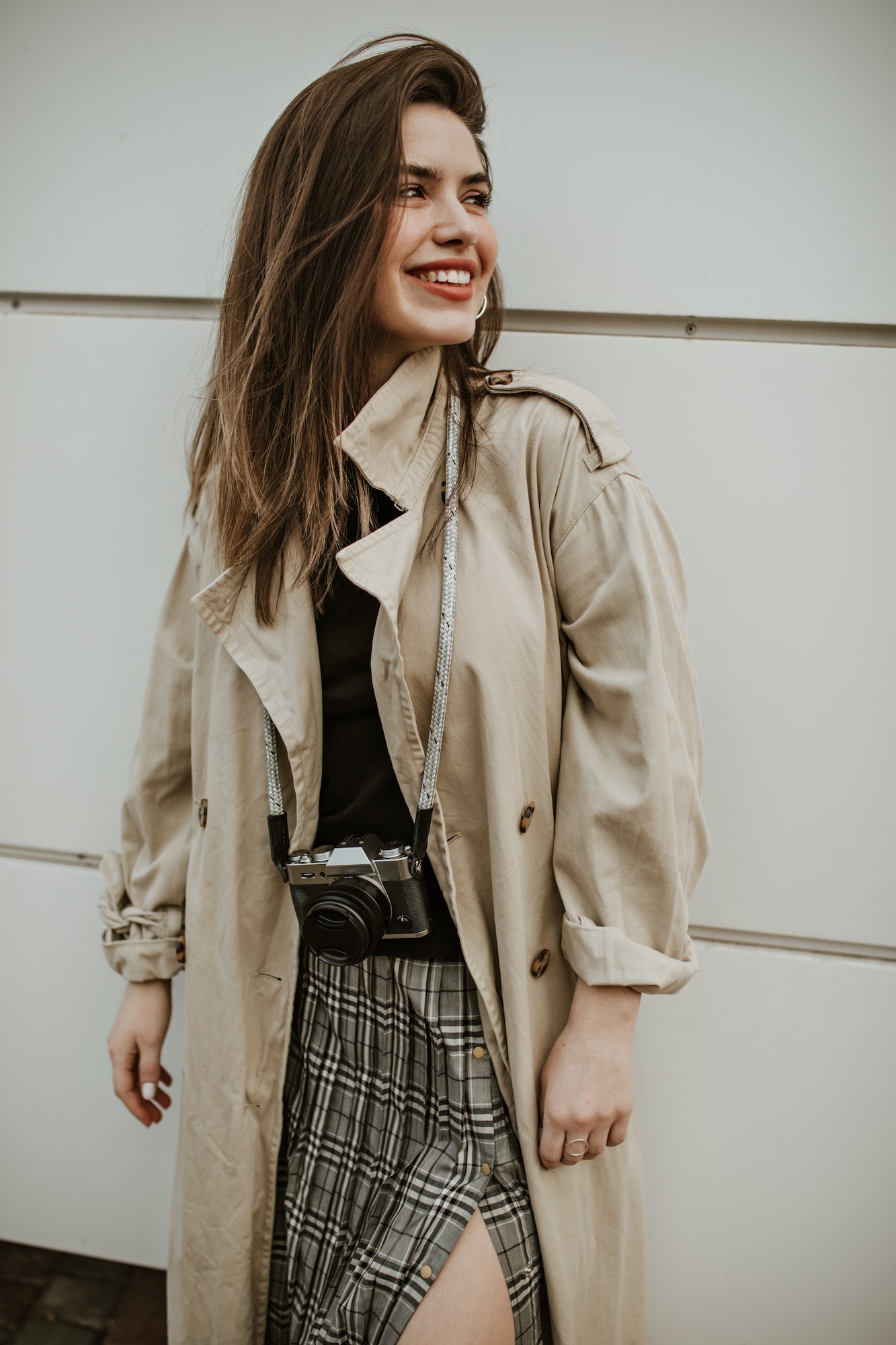 Cheerful attractive woman in checkered skirt and stylish midi beige trench coat smiles, looks away