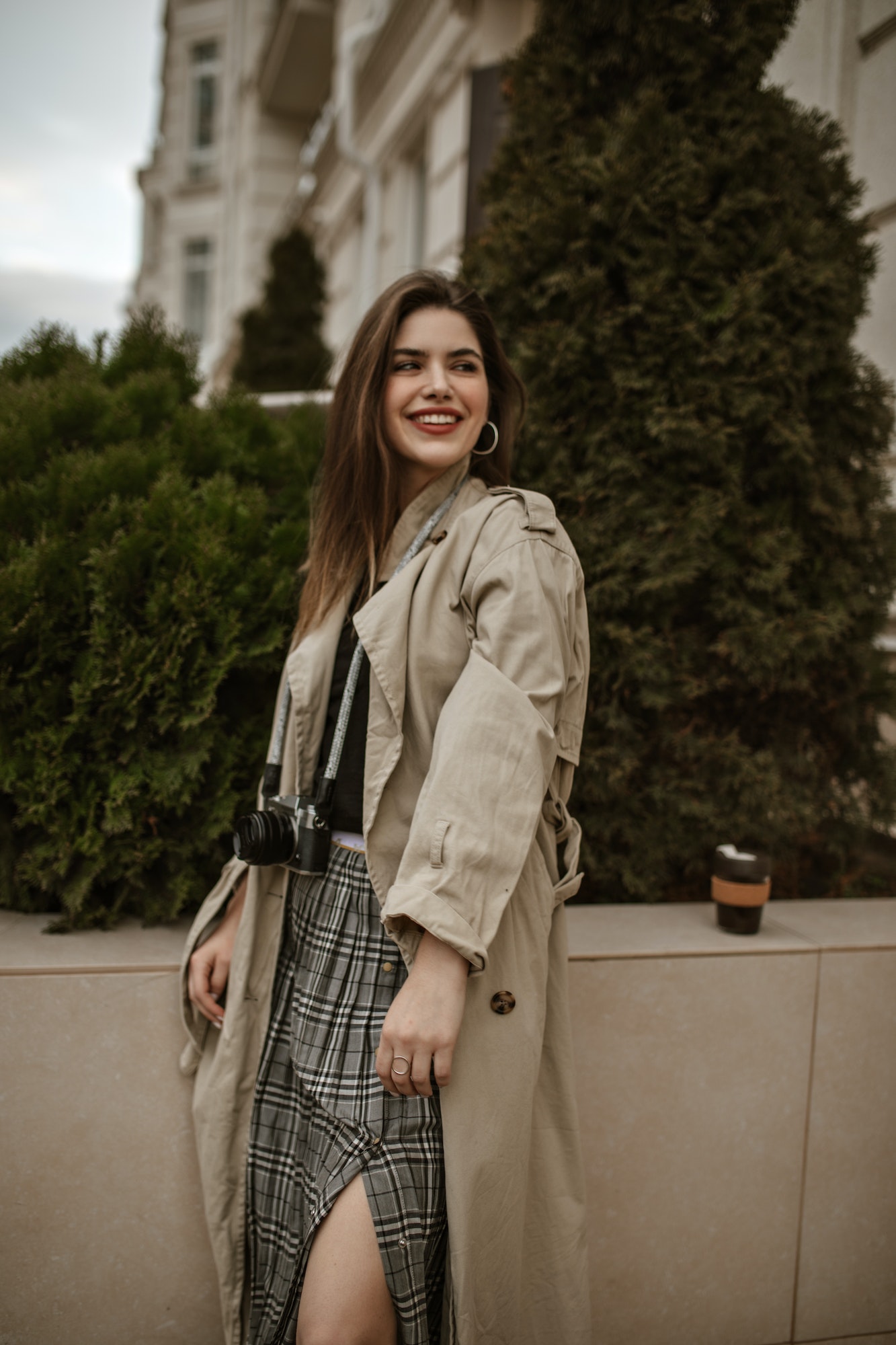 Cheerful brunette young woman in stylish beige trench coat and checkered skirt smilies sincerely an